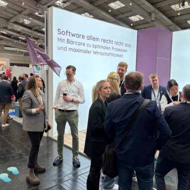 Ein gut besuchter Messestand von Bärcare auf der Pro Care 2026. Im Hintergrund ist eine große, hell leuchtende Wand mit der Aufschrift „Software allein reicht nicht aus. Mit Bärcare zu optimalen Prozessen und maximaler Wirtschaftlichkeit.“ zu sehen. Mehrere Personen in Business-Outfit stehen am Stand im Gespräch zusammen, links hält eine Frau eine lila Flagge mit dem Bärcare-Logo.