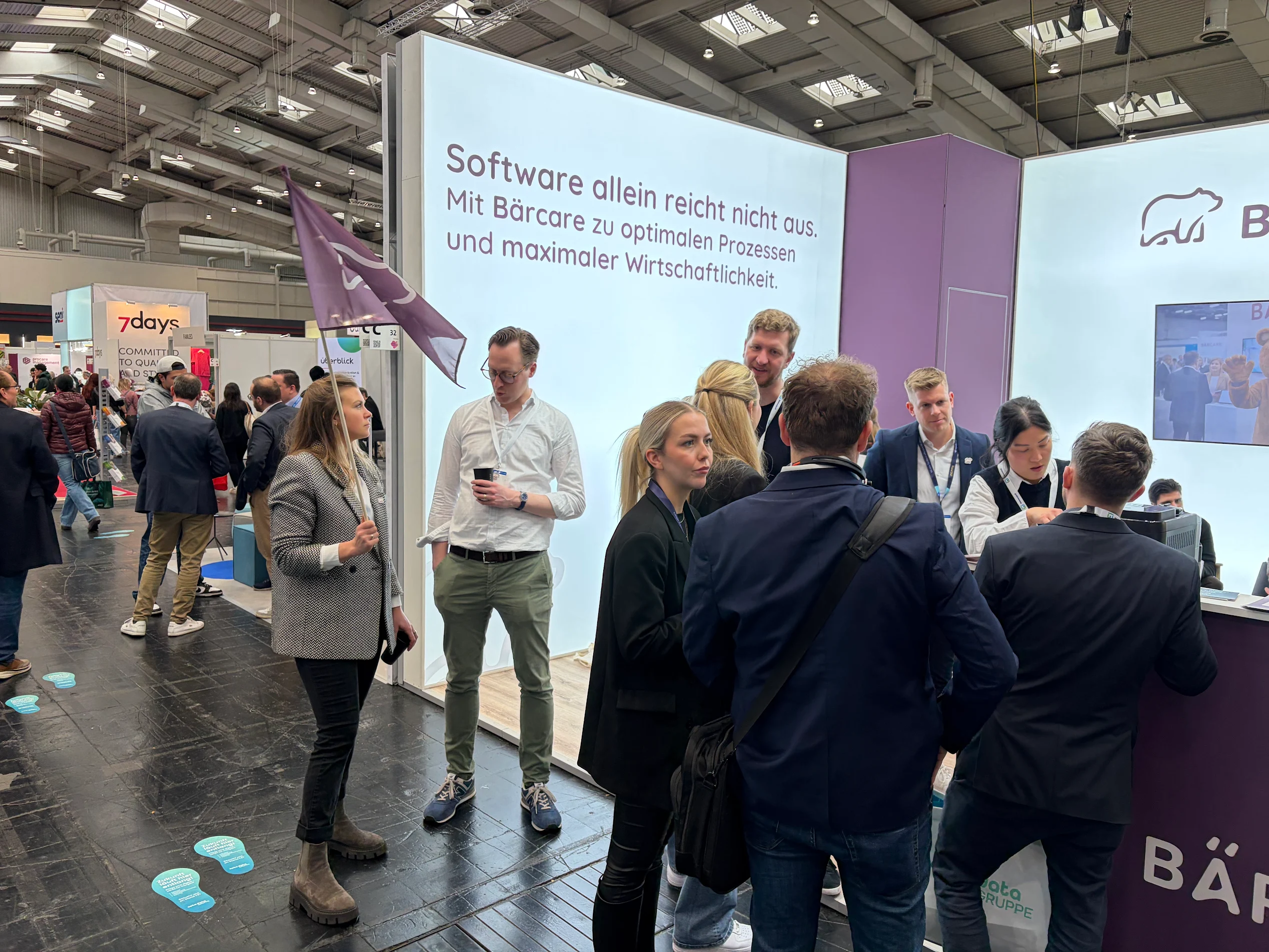 Ein gut besuchter Messestand von Bärcare auf der Pro Care 2026. Im Hintergrund ist eine große, hell leuchtende Wand mit der Aufschrift „Software allein reicht nicht aus. Mit Bärcare zu optimalen Prozessen und maximaler Wirtschaftlichkeit.“ zu sehen. Mehrere Personen in Business-Outfit stehen am Stand im Gespräch zusammen, links hält eine Frau eine lila Flagge mit dem Bärcare-Logo.