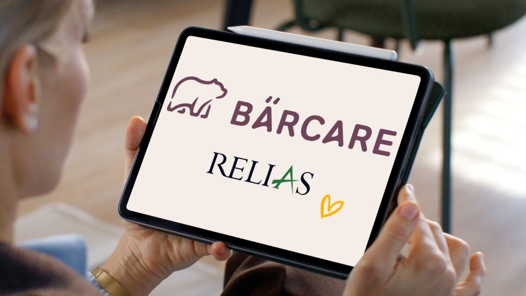 Frau hält ein Tablet in den Händen, auf dem die Logos von Bärcare und Relias mit einem gelben Herzsymbol angezeigt werden