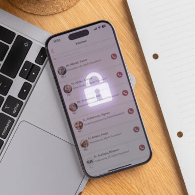 Nahaufnahme eines Smartphones, das auf einer Laptop-Tastatur und einem Holzschreibtisch liegt. Der Bildschirm des Smartphones zeigt eine Liste von 'Klienten' (Kunden oder Patienten) mit Namen, Adressen und Altersangaben, was auf eine Anwendung für das Kunden- oder Pflegemanagement hindeutet. Ein transparentes, leuchtendes Vorhängeschloss-Symbol liegt über der Liste, um die Wichtigkeit des Datenschutzes und der Datensicherheit (möglicherweise im Kontext 'Wem gehören Daten?') zu betonen. Rechts daneben liegen ein Notizblock und ein weißer Stift.