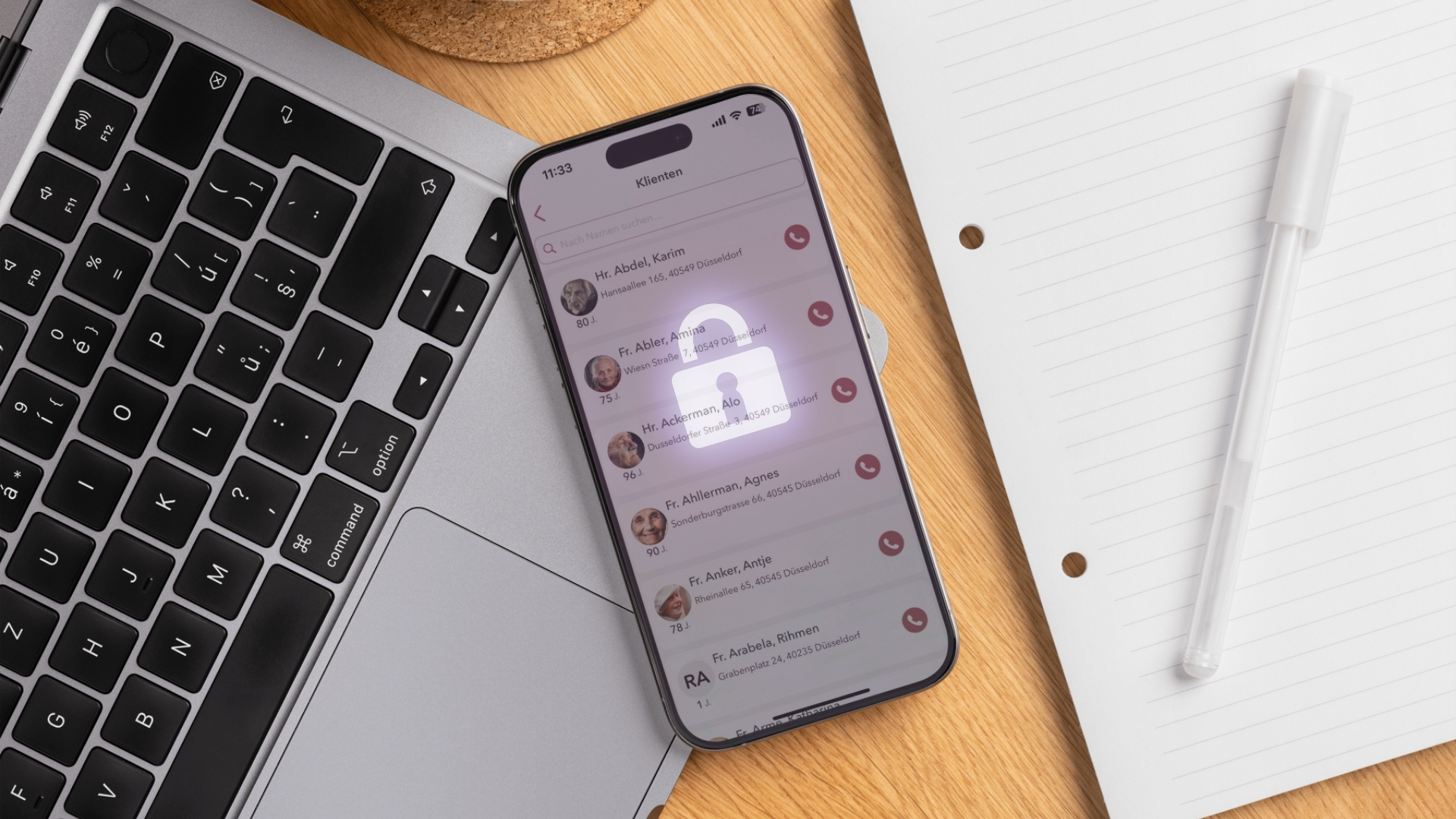 Nahaufnahme eines Smartphones, das auf einer Laptop-Tastatur und einem Holzschreibtisch liegt. Der Bildschirm des Smartphones zeigt eine Liste von 'Klienten' (Kunden oder Patienten) mit Namen, Adressen und Altersangaben, was auf eine Anwendung für das Kunden- oder Pflegemanagement hindeutet. Ein transparentes, leuchtendes Vorhängeschloss-Symbol liegt über der Liste, um die Wichtigkeit des Datenschutzes und der Datensicherheit (möglicherweise im Kontext 'Wem gehören Daten?') zu betonen. Rechts daneben liegen ein Notizblock und ein weißer Stift.