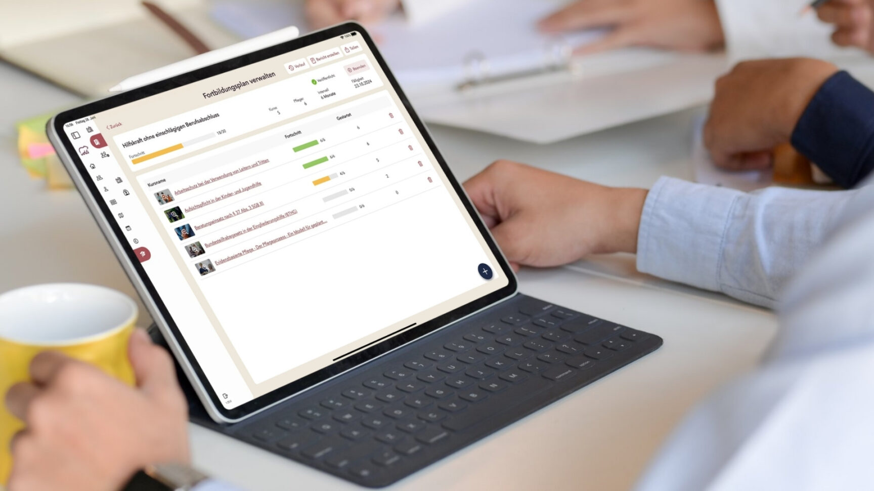 Person arbeitet an einem Tablet mit Tastatur und betrachtet eine Softwareoberfläche zur Verwaltung von Fortbildungsplänen, während weitere Personen im Hintergrund an einem Tisch sitzen.