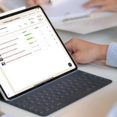 Person arbeitet an einem Tablet mit Tastatur und betrachtet eine Softwareoberfläche zur Verwaltung von Fortbildungsplänen, während weitere Personen im Hintergrund an einem Tisch sitzen.